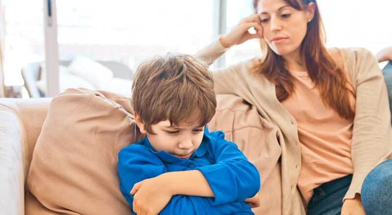A sullen looking young boy with his arms crossed sitting next to his mother on a sofa.