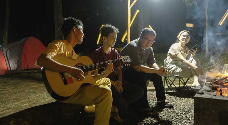 Four young people sitting around a campfire at night as one plays guitar