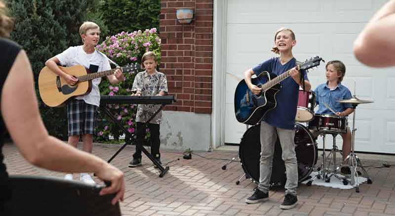 Four young kids performing as a musical band in front of a garage for a couple adults