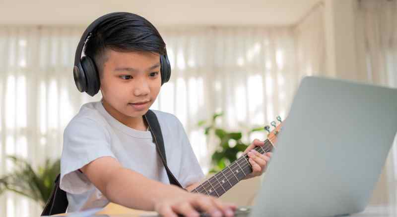 A boy is wearing headphones and using his laptop to assist him with practicing his guitar