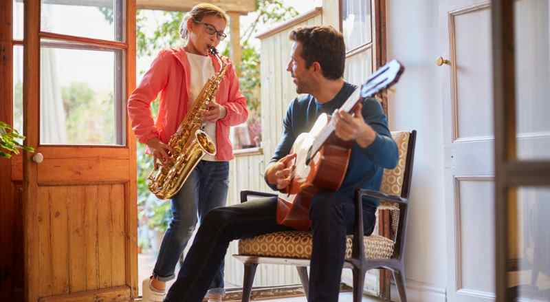 A father and daughter are having fun playing music together.  She is playing the saxophone, and he is playing the guitar